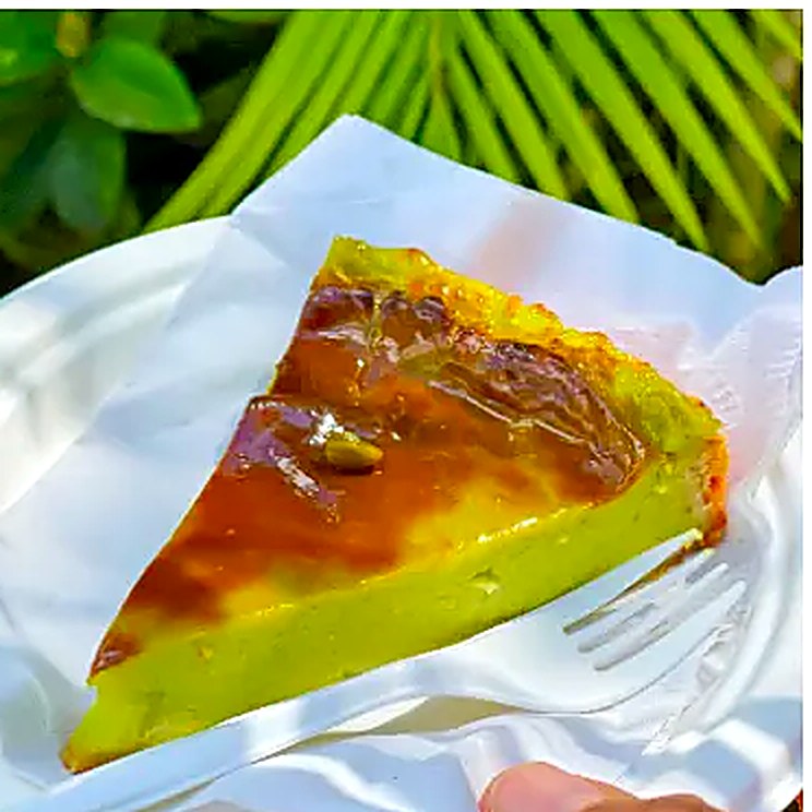 A caramelised slice of pistachio flan on a plate, palm fronds behind